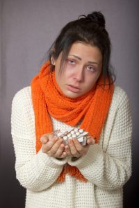 KObieta w białym sweterku i pomarańczowym szaliku, widocznie chora i cierpiąca, trzyma w dłoniacha dużą porcję róznych leków