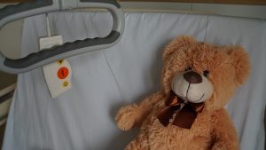 Na łóżku szpitalnym leży brązowy miś z czerwoną kokardką-muszką pod szyją