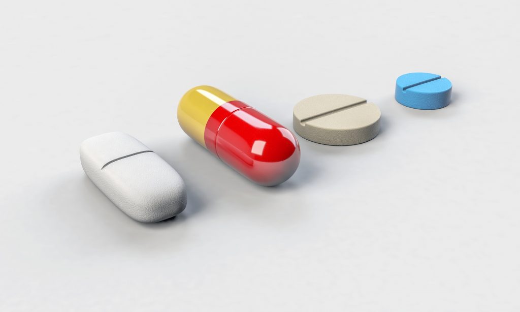 Cztery dawki leków leżące na białym stole: biała podłużba tabletka, czerwono-żółta kapsułka, biała dwudzielna okrągła tabletka i mała, niebieska, okrągła tableteczka
