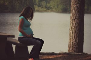 Kobieta w ciąży, w jasnoniebieskiej bluzce z krótkim rękawkiem i spodniach, siedzi na leśnej ławeczce na skraju lasu nad jeziorem. W tle widać pień wysokiego drzewa i spokojną wodę jeziora. Kobieta podrzymuje brzuch opiekuńczym gestem