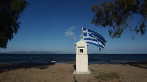 Na plaży, na tle granatowego morza, widoczna mała biała wieżyczka ozdobiona powiewającą na wietrze grecką flagą