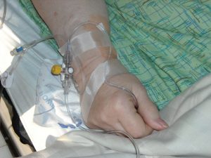 ręka chorej osoby leżącej na łóżku szpitalnym, widoczny wenflon i liczne plastry