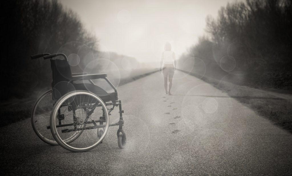 utrzymane w szarościach zdjęcie wózka inwalidzkiego, porzuconego na drodze, w oddali półprzeźroczysta osoba odchodząca w kierunku horyzontu