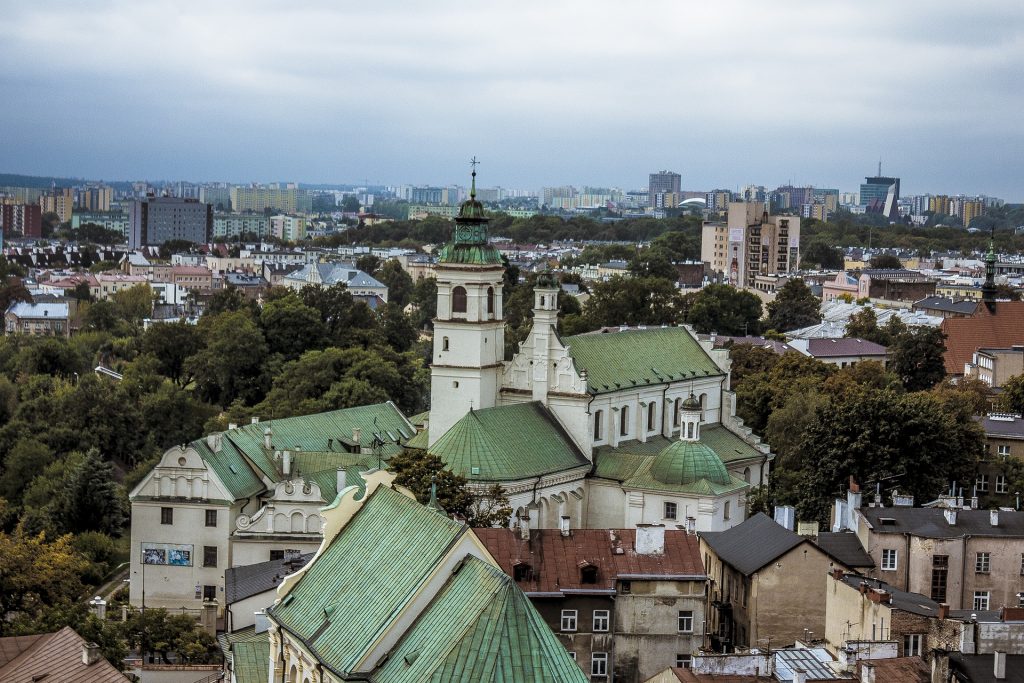 Dachy miasta Lublina z lotu ptaka, na pierwszym planie budynki sakralne o zielonych dachach, w tle inne budynki i dużo zieleni, niebieskie niebo z chmurami