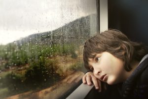 Smutny chłopiec w wieku około 10-12 lat oparty o okno, patrzy w dal przez pokrytą kroplami deszczu szybę