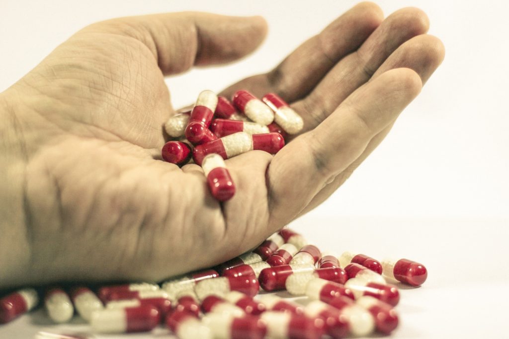 Na białym tle widoczna dłoń trzymająca dużą ilość biało-czerwonych kapsułek leków