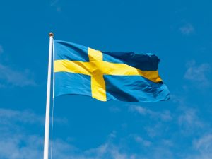 Szwedzka niebieska flaga z żółtym krzyżem na białym maszcie powiewająca na tle niebieskiego nieba