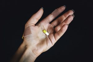 Na ciemnym tle biała ręka delikatnej budowy z długimi paznokciami i plastrami na palcach, z umieszczoną biało-żółtą kapsułką na dłoni