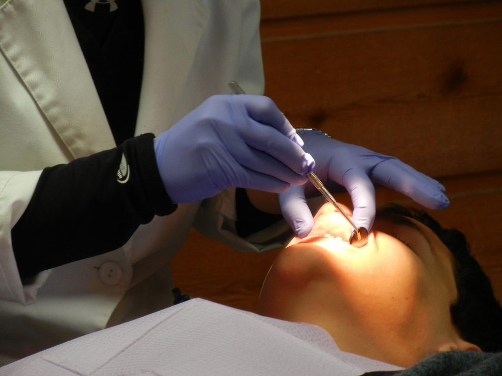 Pacjent u stomatologa: widoczna jest głowa od strony podbródka i oświetlona pole jamy ustnej oraz ręce lekarza ubranego w biały fartuch i niebieskie rękawiczki
