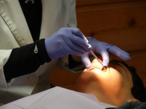 Pacjent u stomatologa: widoczna jest głowa od strony podbródka i oświetlona pole jamy ustnej oraz ręce lekarza ubranego w biały fartuch i niebieskie rękawiczki