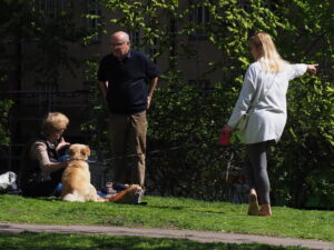 Dwie osoby stojące na zielonej trawie i jedna siedząca. Siedząca obejmuje żółtawego psiaka.