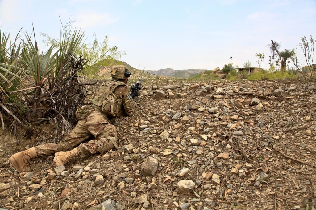 Amerykański żołnierz leżący na żwirowym podłożu pod krzakiem, celujący w nieznany cel poza horyzontem