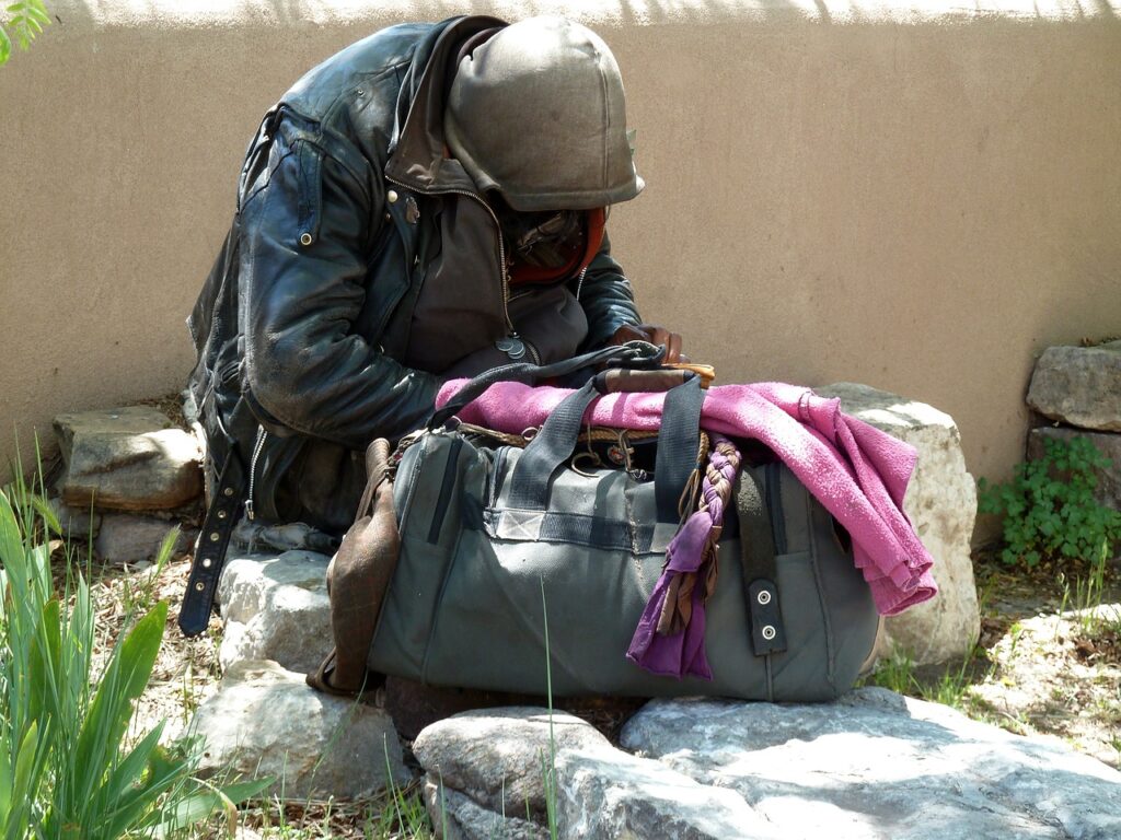 Osoba w kryzysie bezdomności, siedząca pod ścianą, przed nią torba z dobytkiem